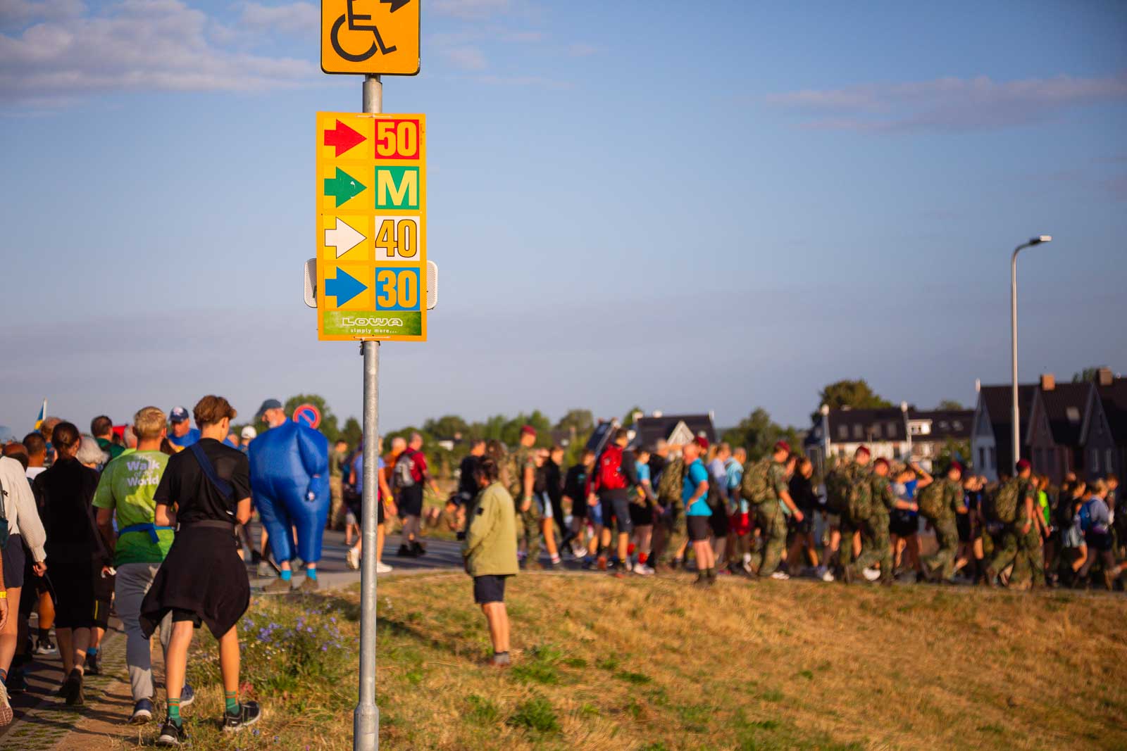 Wegwijzer met gekleurde pijlen voor de 30, 40 en 50 km-routes van de 4Daagse, terwijl wandelaars op de achtergrond voorbij trekken.