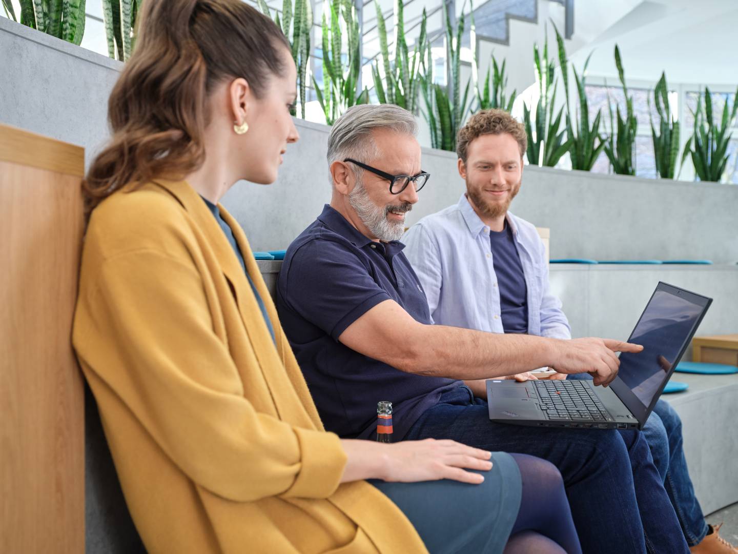 Drie personen zitten in een moderne, vergroende werkomgeving. Een man wijst naar het scherm van een laptop, terwijl de anderen aandachtig luisteren. De scène straalt een ontspannen en productieve sfeer uit.