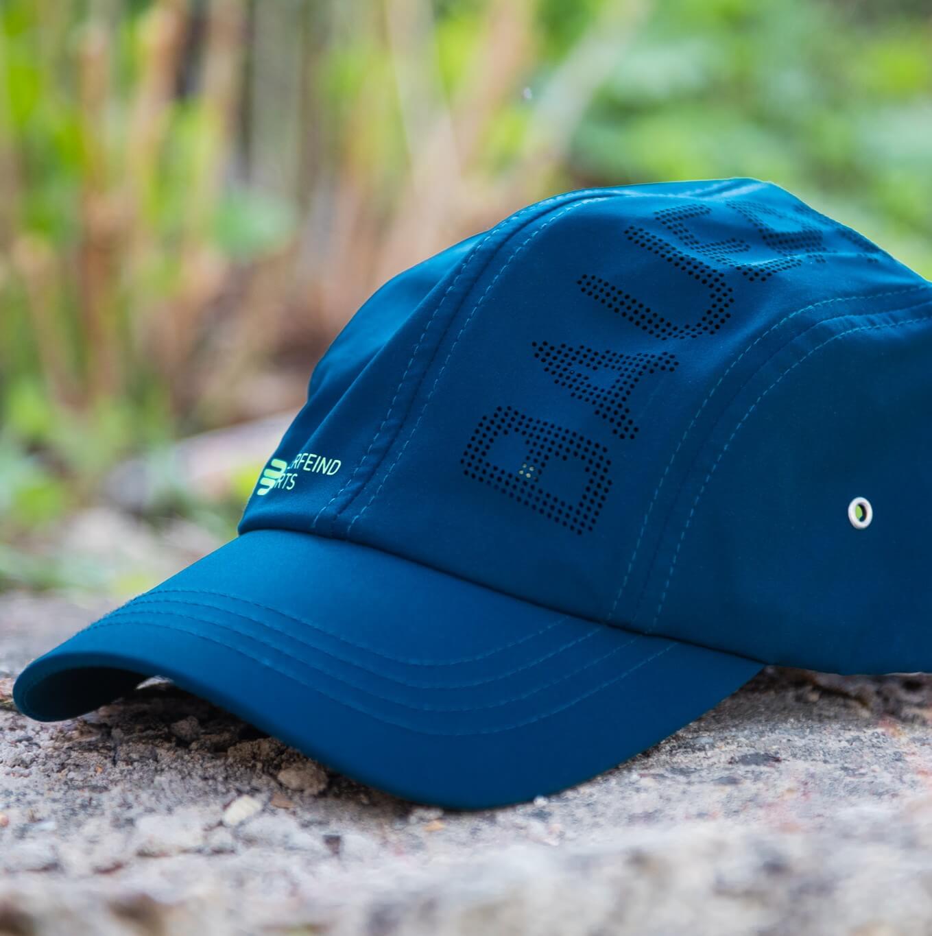 Blauwe Bauerfeind Sports pet in zijaanzicht op een stenen ondergrond, met gebogen klep, groen merklogo en geperforeerde BAUERFEIND-tekst op het paneel – ideaal voor frisse momenten buiten.