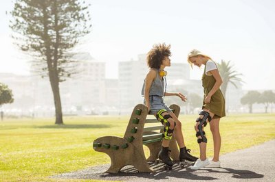 De afbeelding toont twee vrouwen in gesprek bij een parkbank. Beiden dragen knieorthesen van Bauerfeind om hun geblesseerde knie te stabiliseren.