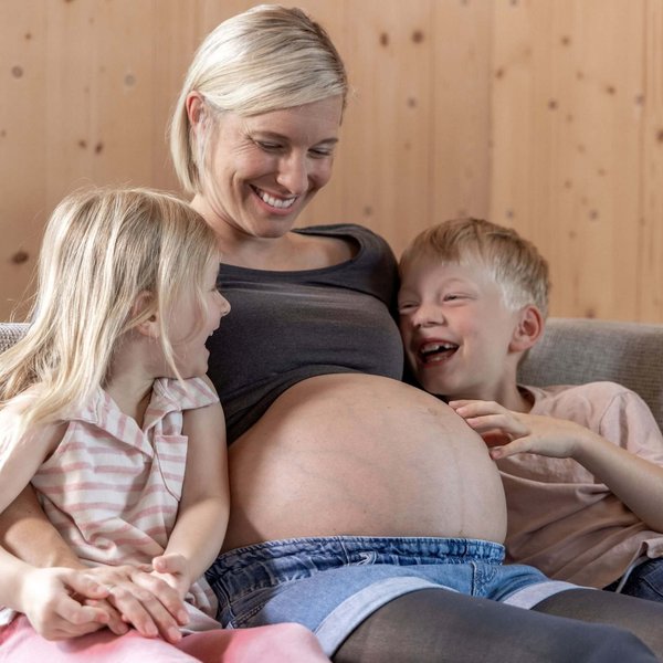 Zwangere vrouw zit op een bank en lacht terwijl haar twee kinderen vrolijk haar babybuik aanraken.