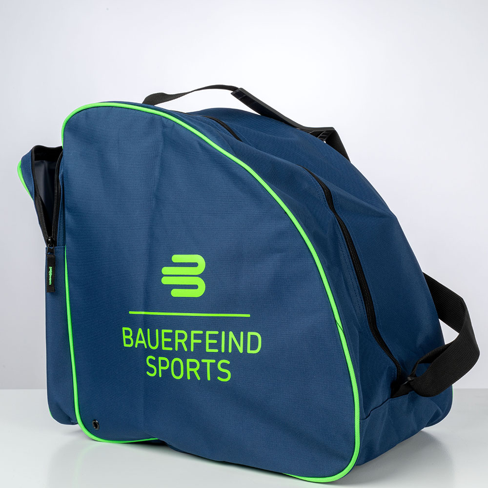 De Bauerfeind Sports skitas staat buiten op een houten bank. De tas is blauw met een groene rand en heeft een zwarte draagriem. De bomen op de achtergrond suggereren een buitenomgeving.