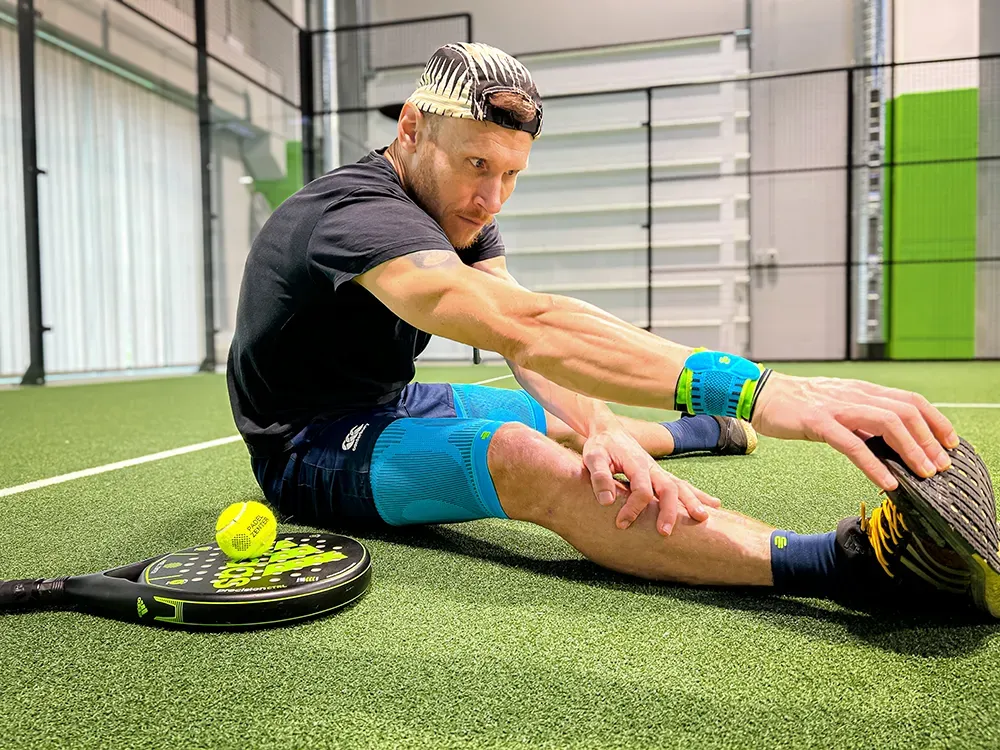 Een sporter zit op de grond van een padelbaan, rekt zijn beenspieren en draagt compressieproducten aan arm, been en pols. Naast hem liggen een padelracket en een bal.