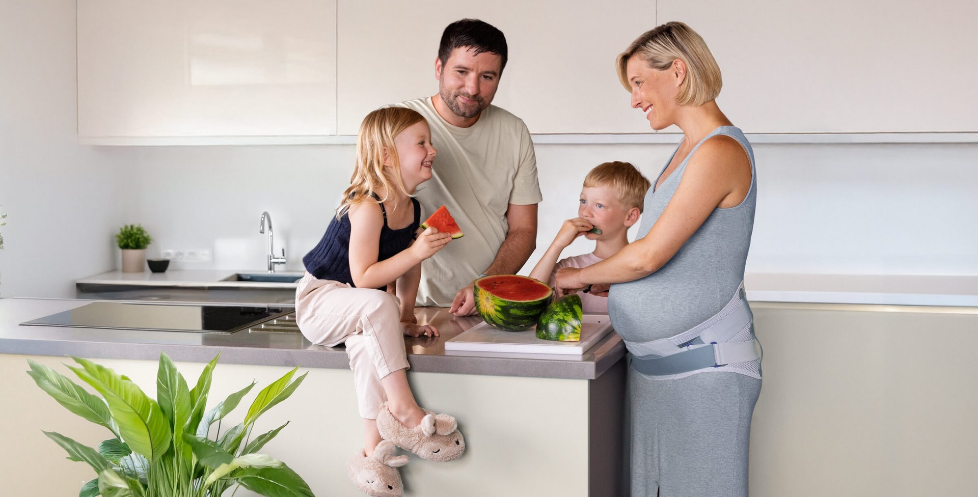 Een gezin staat in een moderne keuken rond een kookeiland. Een meisje zit op het werkblad met een stuk watermeloen, een jongen eet meloen, en een zwangere vrouw in een grijze jurk met buiksteun snijdt de watermeloen. Iedereen lacht.