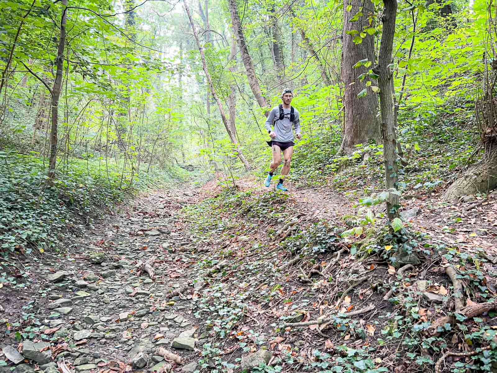 Een trailrunner rent op een rotsachtig pad door een dicht bos. De omgeving is groen en rustig, ideaal voor intensieve buitentraining.