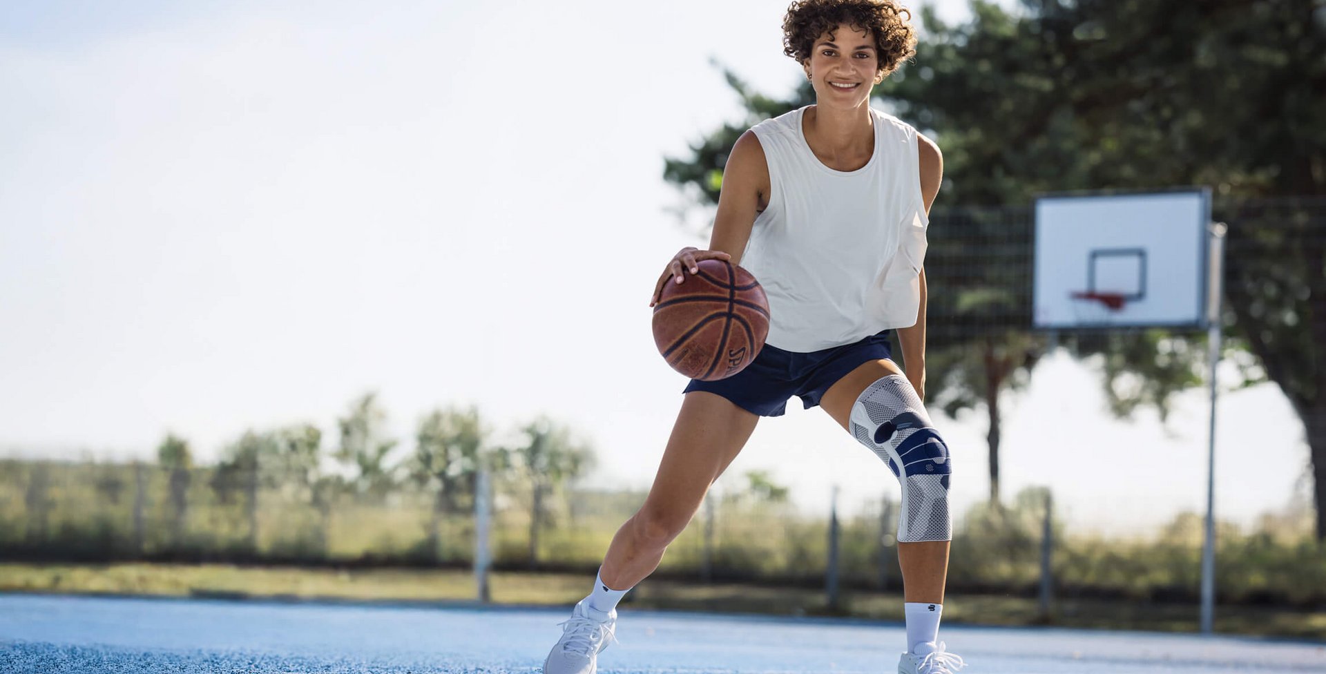 Jonge vrouw met wit hemdje, donkerblauwe sportbroek en witte kniebandage speelt basketbal op een buitenveld met blauwe ondergrond; ze dribbelt de bal en lacht naar de camera, op de achtergrond zijn een basketbalring en bomen te zien.