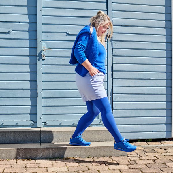 Vrouw met blond haar en blauwe kleding, waaronder een blauwe panty, trui en sneakers, loopt een klein betonnen trapje af voor een lichtblauwe houten wand met rode rand.