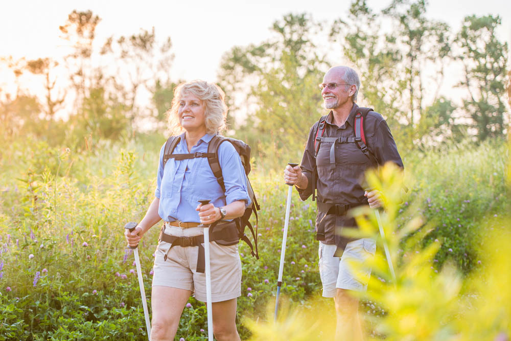 Een ouder stel wandelt met trekkingstokken door een bloeiende zomerse weide in het warme avondlicht. Beiden dragen lichte wandelkleding en rugzakken en zien er tevreden uit. De scène straalt rust, vitaliteit en wandelplezier uit.