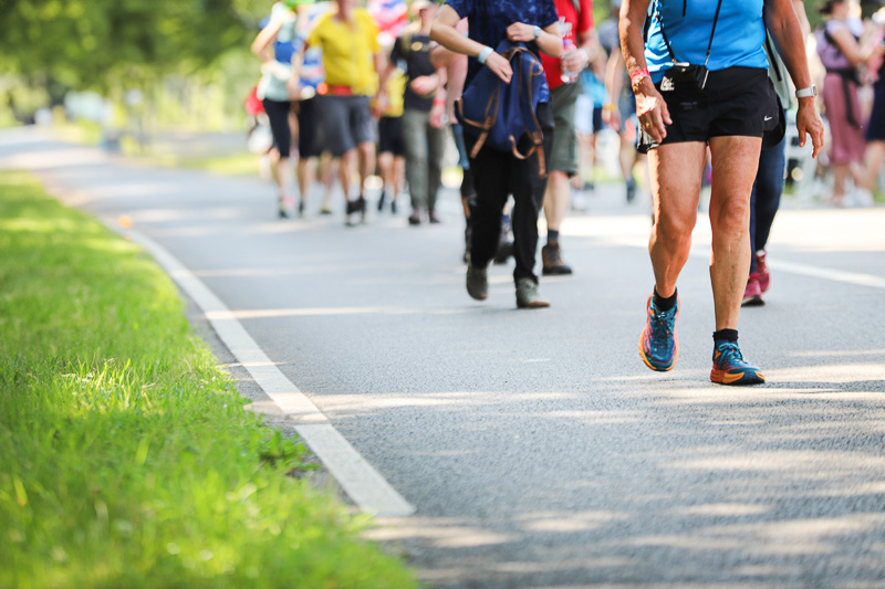 Een groep wandelaars loopt over een afgesloten weg in een groene omgeving op een zonnige dag. Op de voorgrond is een man in blauwe sportkleding en hardloopschoenen te zien.