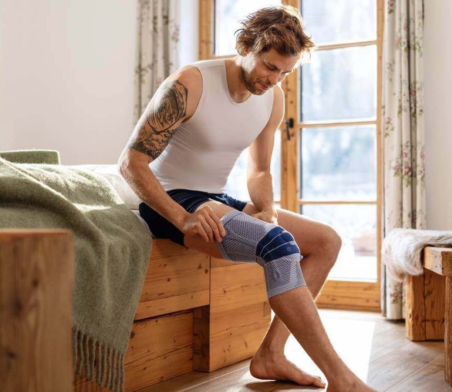 Een jonge man zit op een bed en doet een bandage aan. Het gaat om de GenuTrain P3 van Bauerfeind, die wordt gebruikt bij de behandeling van kniepijn.