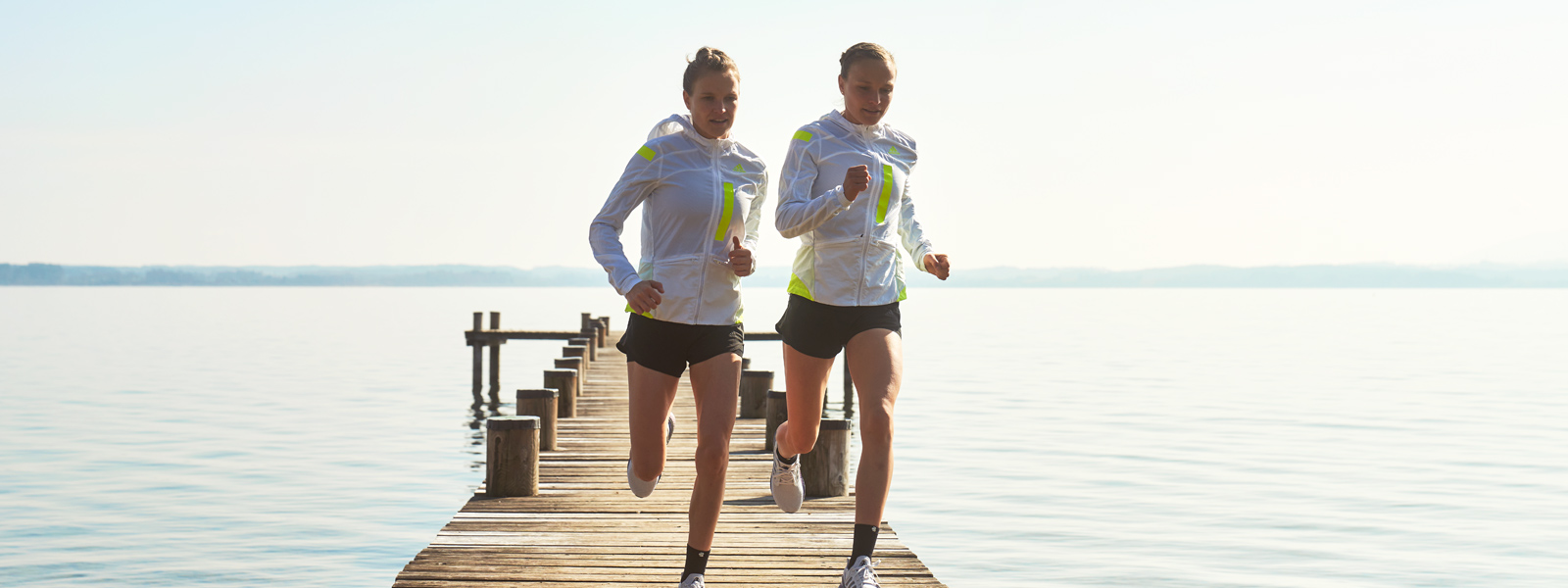 Twee lopers in witte hardloopjassen en met korte broek rennen over een steiger naar de bank