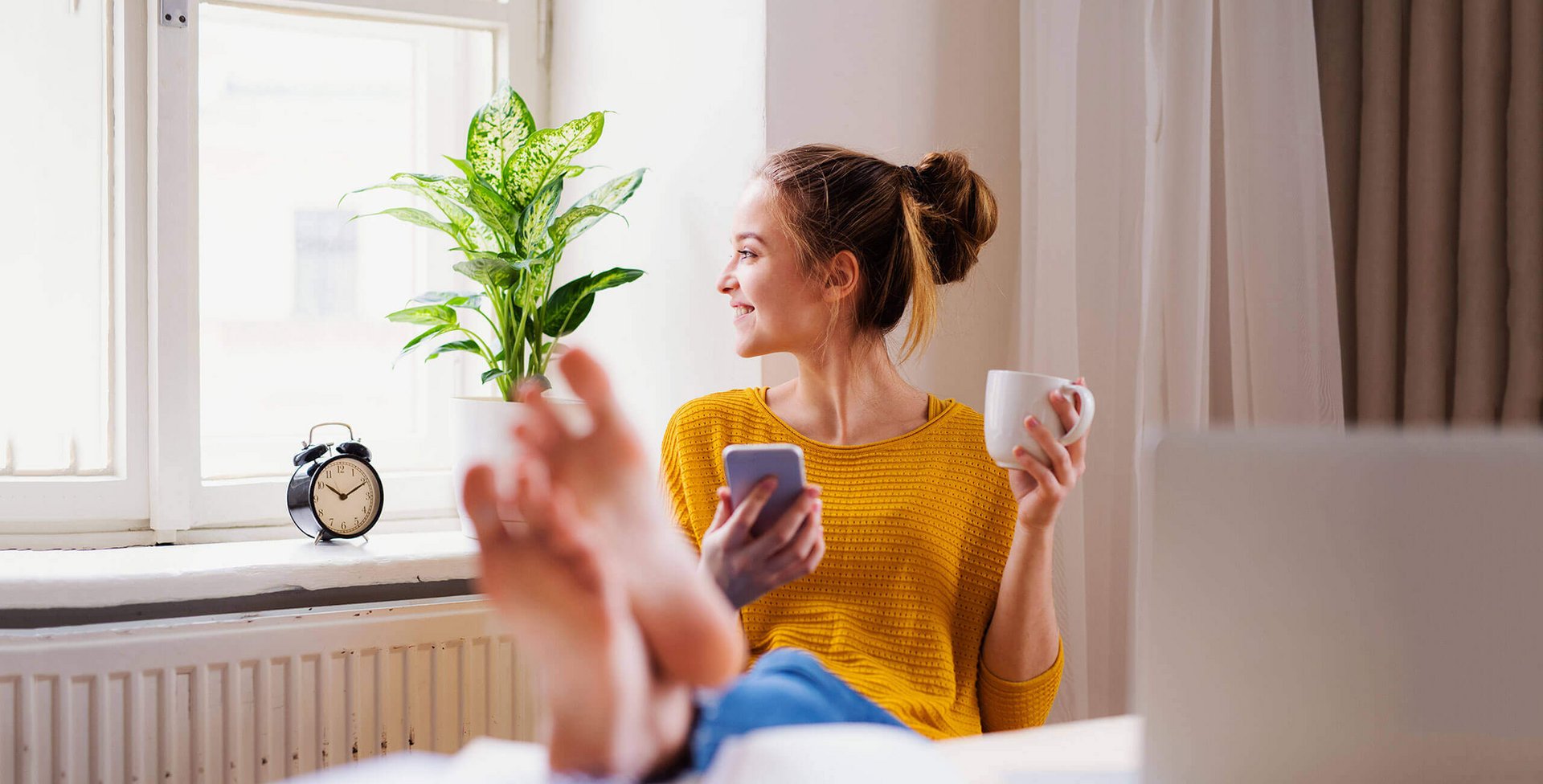 Op de foto zit een jonge vrouw op een stoel met haar voeten op de tafel voor zich. In haar linkerhand houdt ze een kopje, in haar rechterhand een smartphone. Regelmatige training van de voetspieren kan pijn voorkomen.