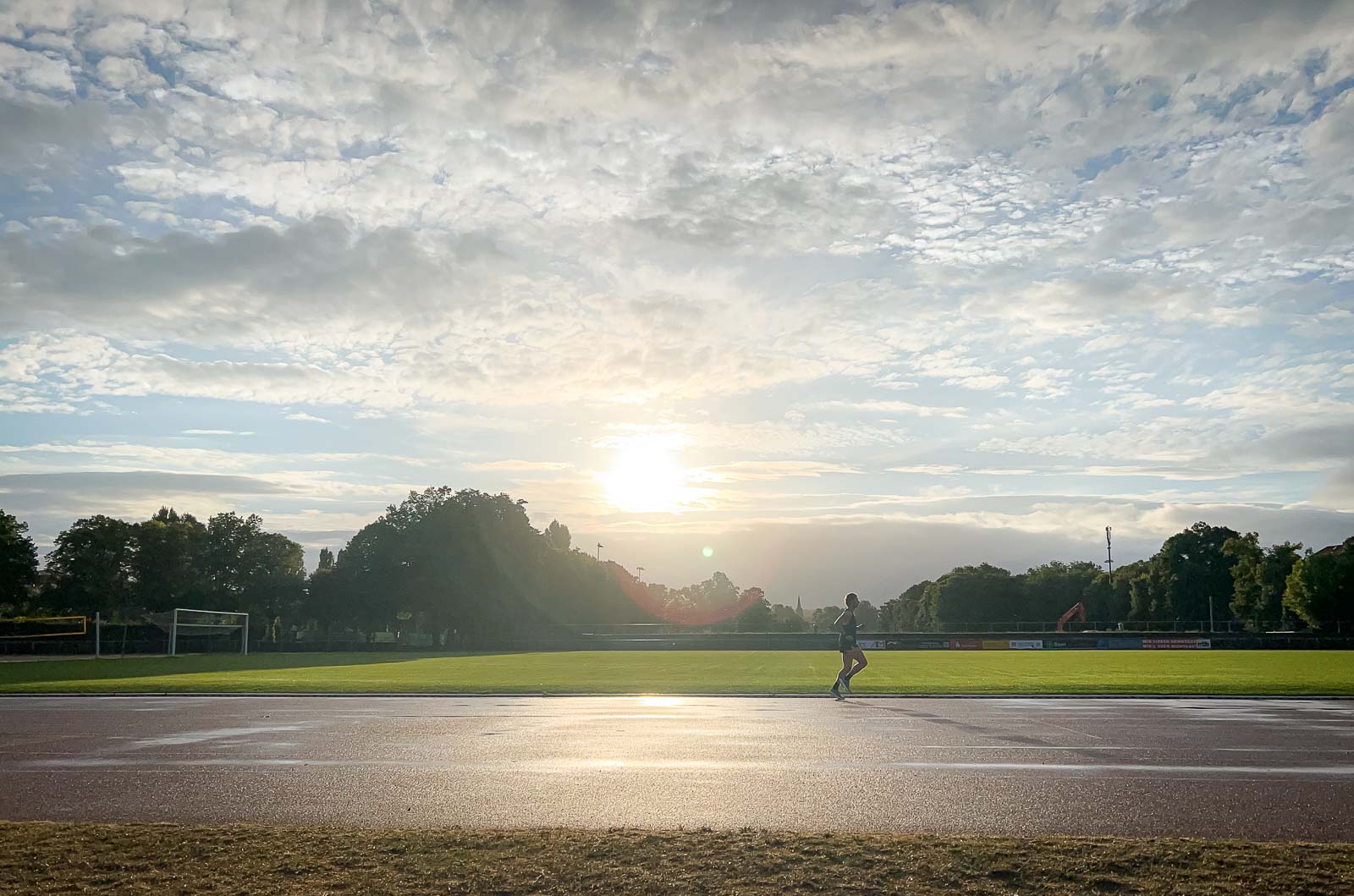 Een loper op een natte renbaan bij zonsopgang. De lucht is bewolkt, maar de zon breekt door terwijl de loper zijn ronde afmaakt.