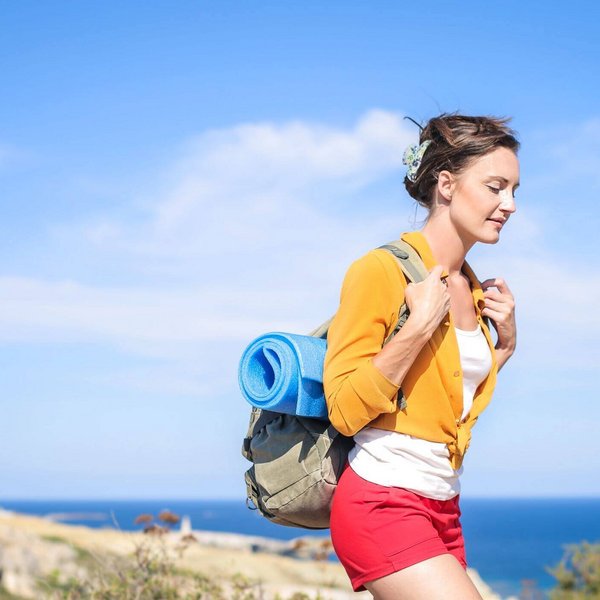 De afbeelding toont een jonge vrouw met een wandelrugzak in de zon aan zee. In de warme zomermaanden valt het ons meestal gemakkelijker om actief te worden en bewegingsgebrek tegen te gaan.