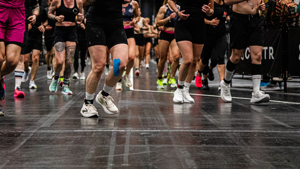 Dynamische close-up van de benen van meerdere atleten die over een donkere indoor hardloopbaan lopen tijdens een HYROX-wedstrijd; sportschoenen, wedstrijdkleding en blauwe kinesiotape op een been tonen de intensieve beweging