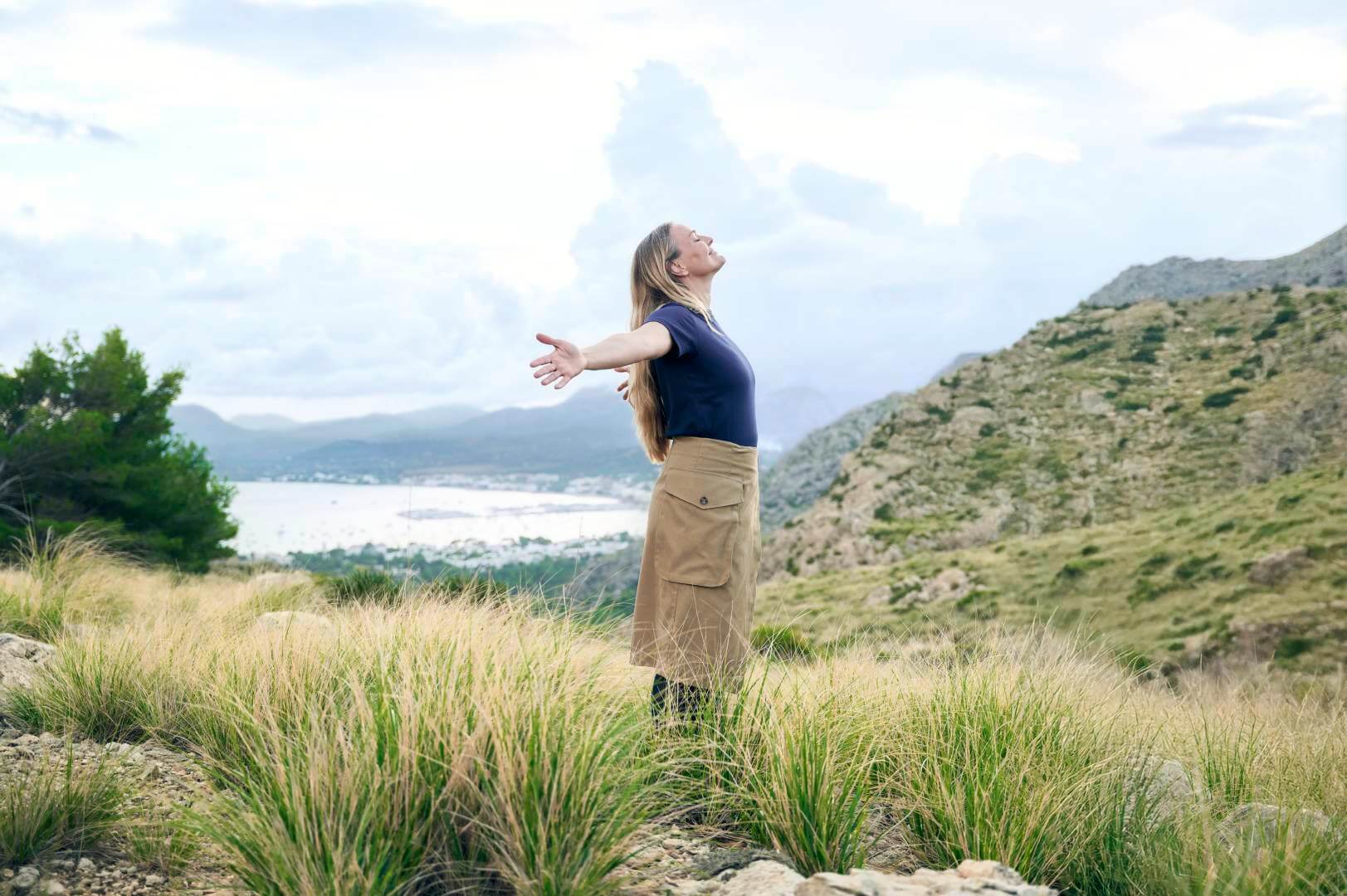 Een vrouw met lang los haar staat op een heuvel met uitzicht op een kustlandschap. Met gespreide armen geniet ze van de frisse lucht; bij haar outfit in aardtinten draagt ze subtiele VenoTrain compressiekousen.