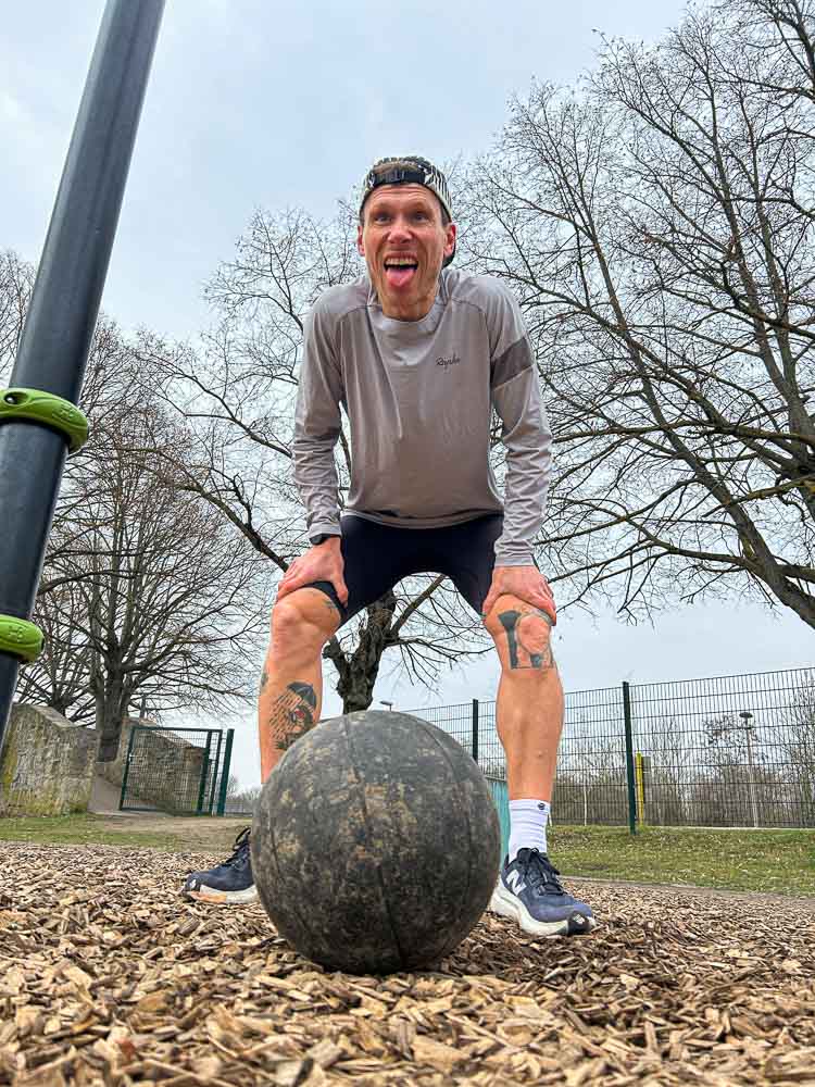 Een sporter staat na een intensieve sessie in een outdoor trainingspark met de handen op de knieën boven een medicine ball. Het beeld laat vermoeidheid zien na een functionele workout die past bij HYROX-training zonder sportschool.