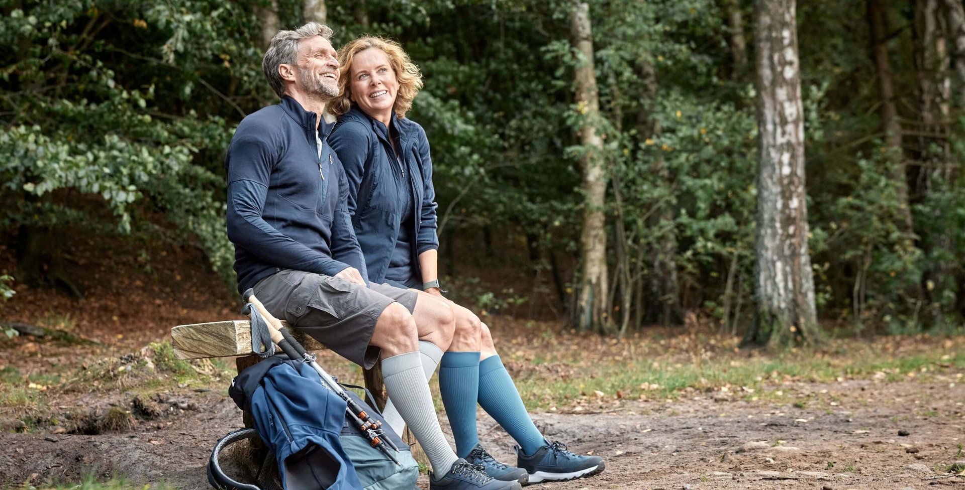 Een lachend stel zit op een houten bank in het bos en draagt stevige VenoTrain hero compressiekousen in grijs en blauw. Naast de bank ligt een wandelrugzak met wandelstokken.
