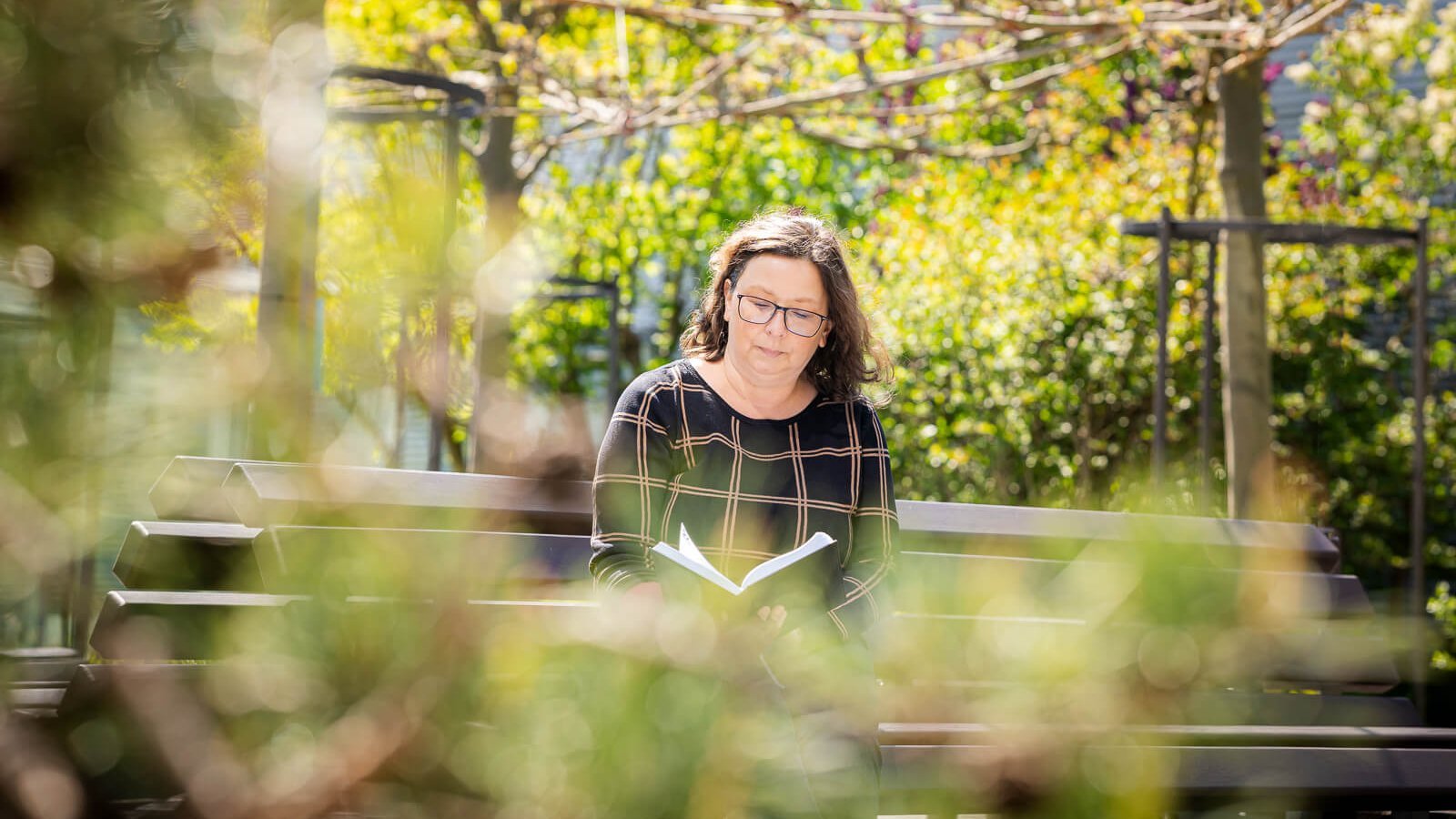 Katrin, een lymfoedeempatiënte, zit op een bank in een zonnige tuin en leest een boek. Om haar heen staan bomen en planten die het beeld met een natuurlijke en vredige sfeer omgeven.