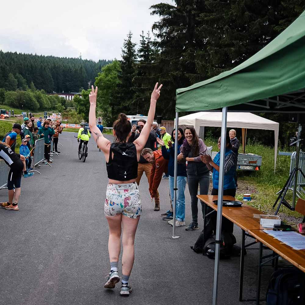Een vrouw in sportkleding rent door de Rennsteig en heft beide armen om het vredesteken te maken terwijl de toeschouwers haar aanmoedigen.