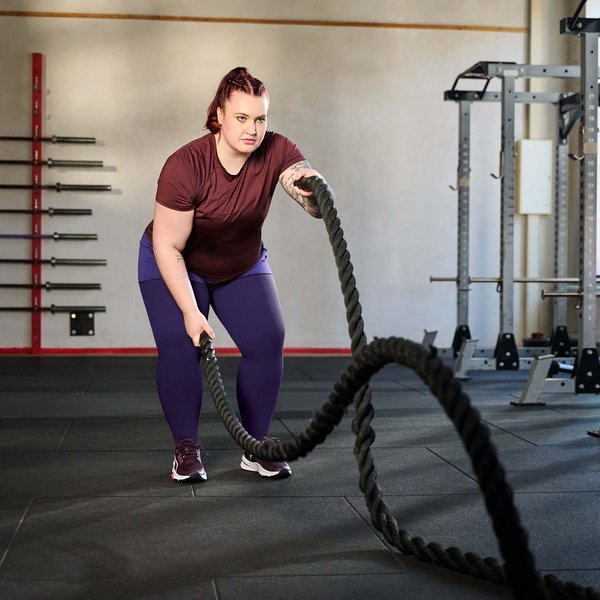 Een vrouw traint in een sportschool met battle ropes. Ze draagt een bordeauxrood T-shirt, een paarse legging en sportschoenen. Op de achtergrond zijn halters aan de wand en krachttoestellen te zien.