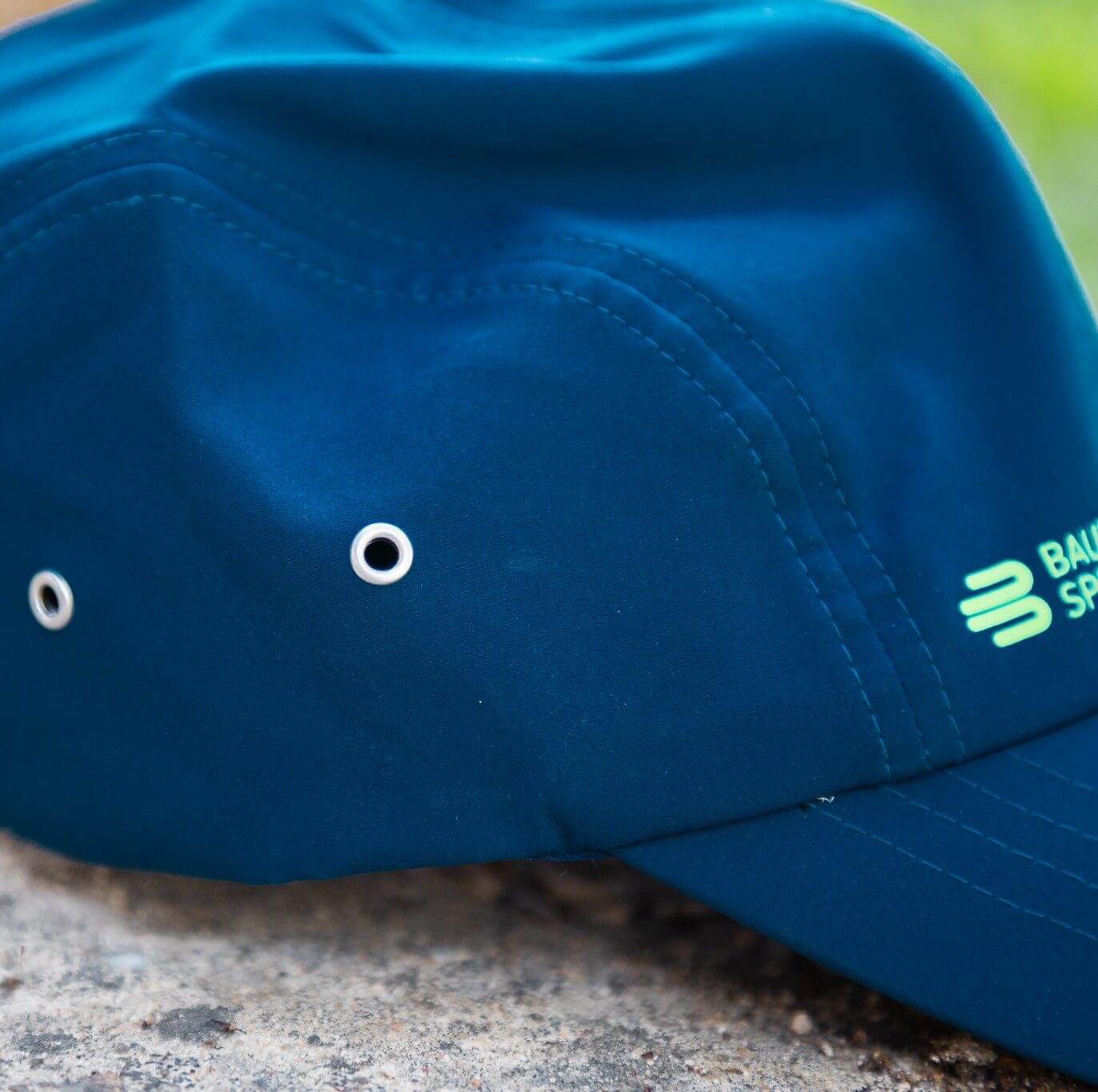 Detailopname van de blauwe Bauerfeind Sports pet met metalen oogjes aan de zijkant, nauwkeurige stiksels en een gedeeltelijk zichtbaar groen logo op het frontpaneel – een sportief accessoire voor training, ritten en dagelijks gebruik.