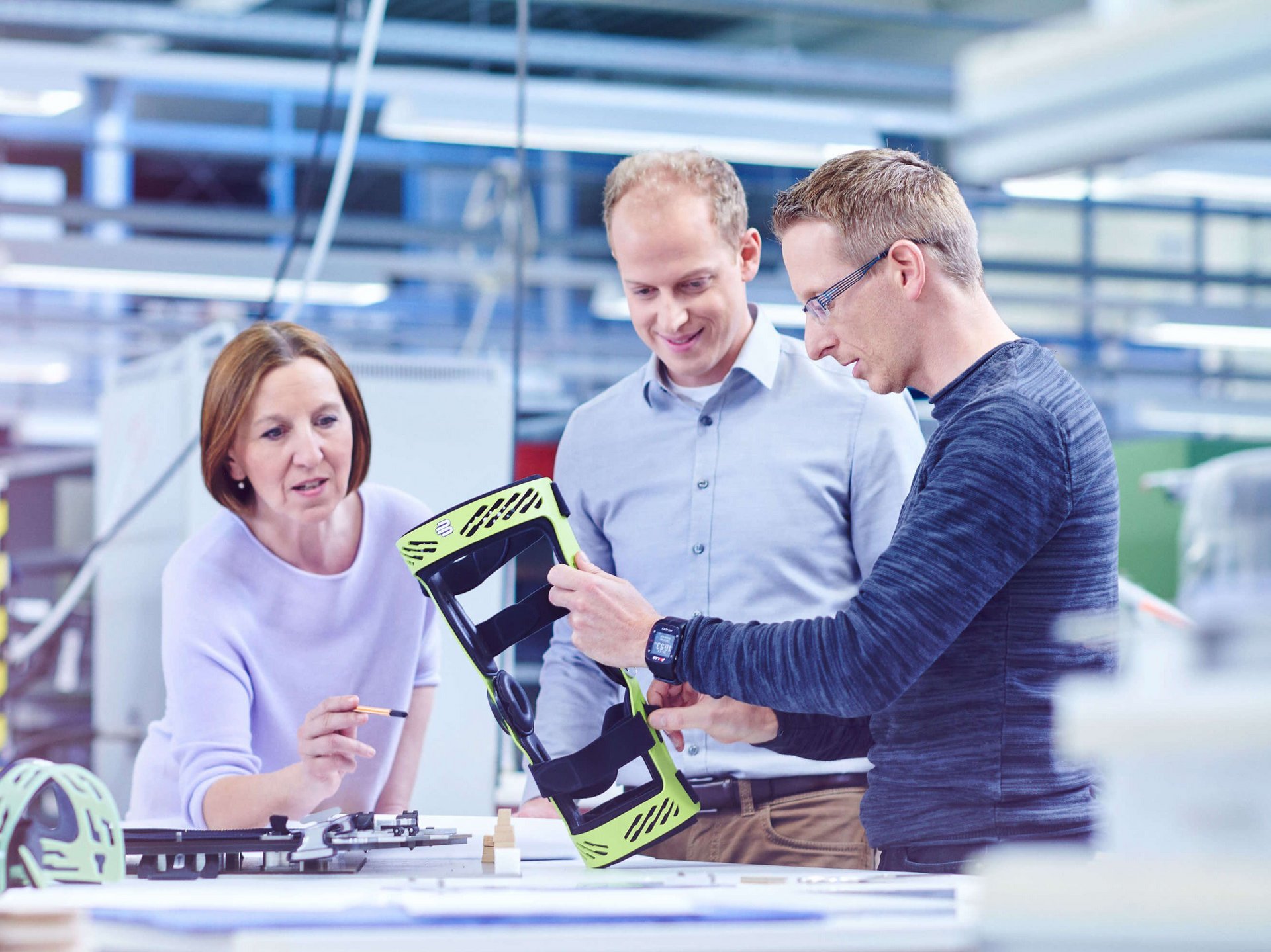 Drie personen staan aan een grote tafel in de ontwikkelingsafdeling en kijken naar de frame-orthese SecuTec Genu. Ze bekijken en bespreken een lichtgroen technisch onderdeel, waarschijnlijk een behuizing of houder, dat de man rechts in zijn handen houdt.