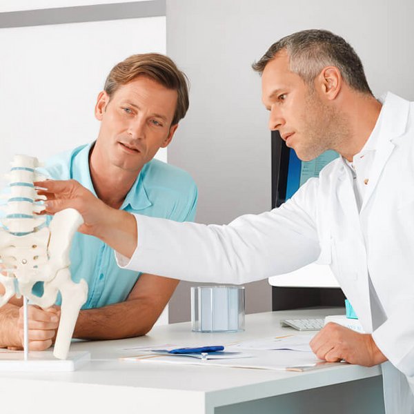 Een arts in een witte jas legt een mannelijke patiënt met behulp van een model van de menselijke wervelkolom en het bekken een medische diagnose uit. De patiënt luistert aandachtig en beiden bevinden zich in een moderne behandelkamer.