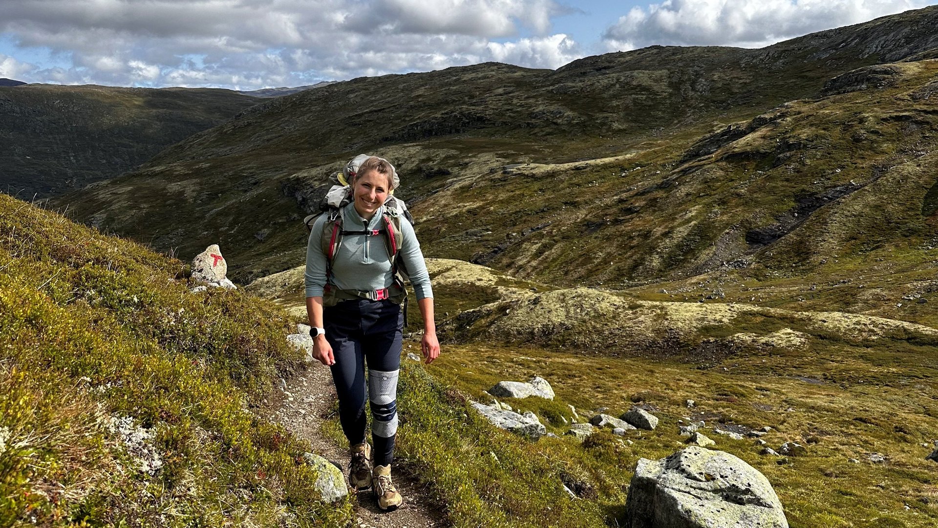 Antonia Horn tijdens een wandeling in een indrukwekkend berglandschap. Ze draagt een GenuTrain-kniebandage en wandelt over een smal pad door de ongerepte omgeving. De hemel is deels bewolkt, het uitzicht biedt groene heuvels en rotsachtige stukken.