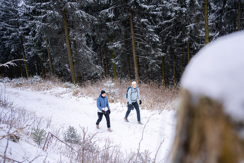 wee vrouwen wandelen door een besneeuwd boslandschap. Ze dragen warme winterkleding en gebruiken wandelstokken terwijl ze een pad volgen. Op de achtergrond zijn met sneeuw bedekte bomen en een dicht bos te zien.