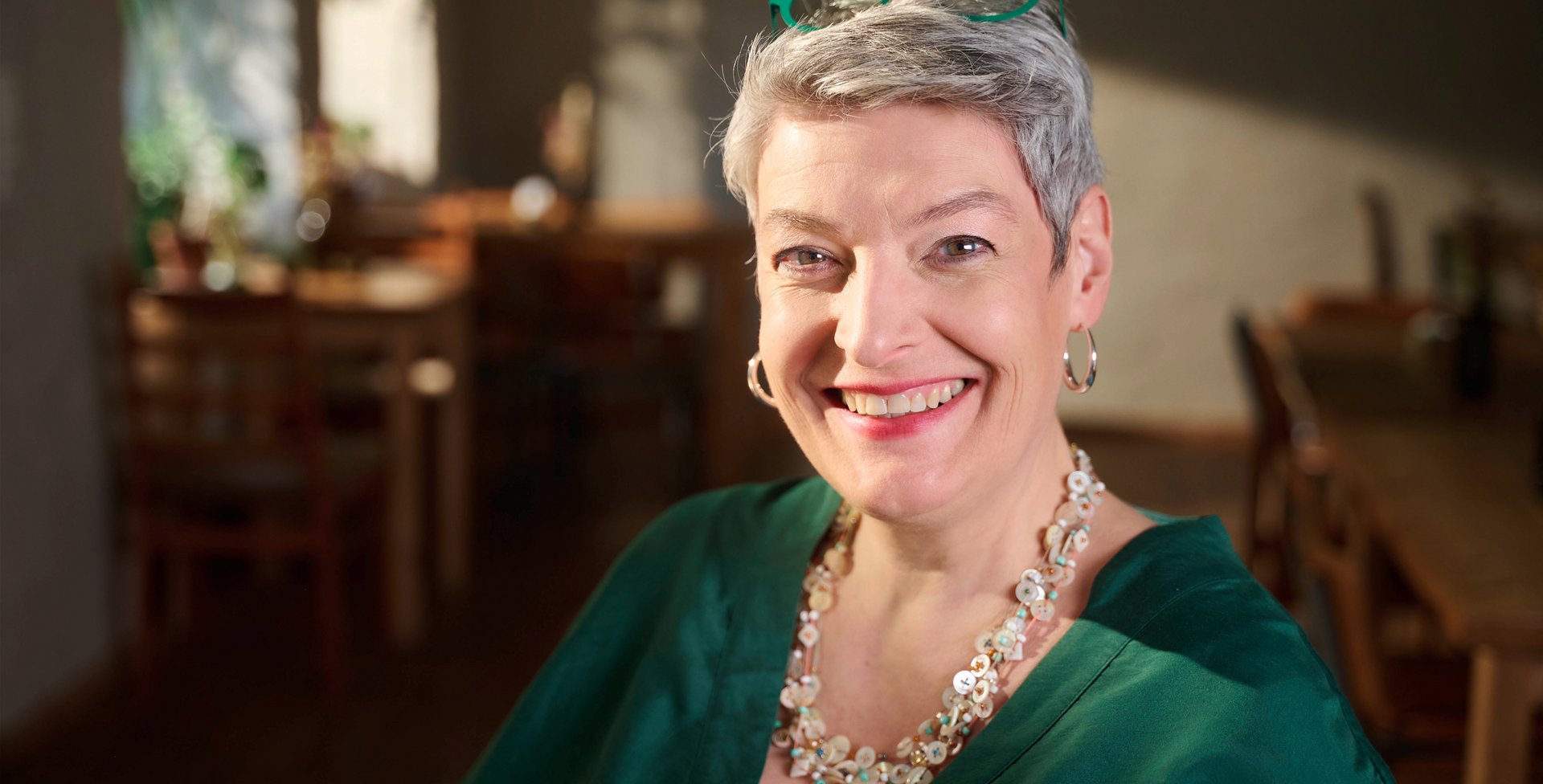 Portret van een lachende vrouw met kort grijs haar, een groene bril op haar hoofd, een groene blouse en een opvallende ketting, zittend in een gezellig café met houten meubels op de achtergrond.