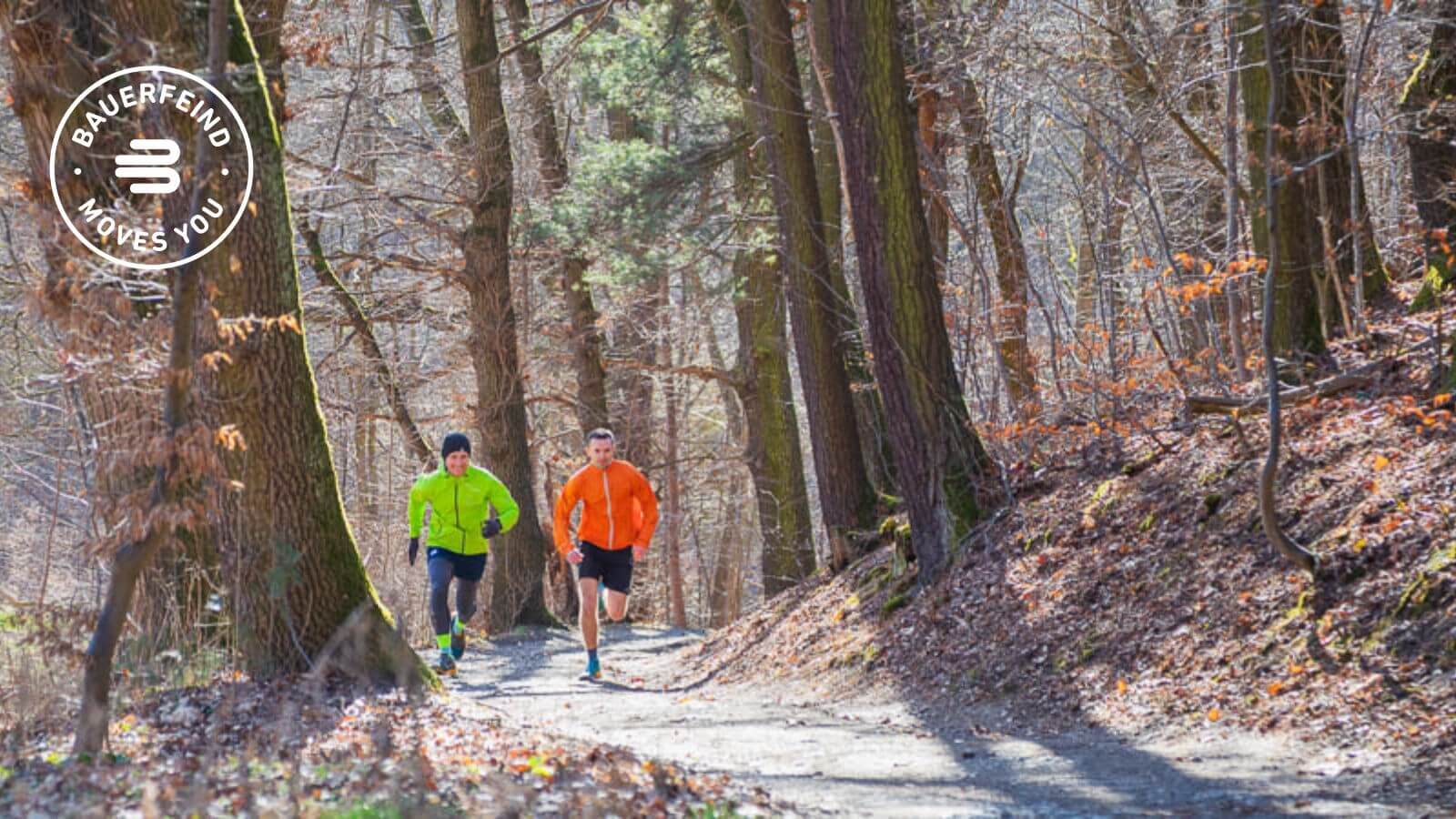 Twee hardlopers in felgekleurde jassen (groen en oranje) joggen over een zonnig bospad, omringd door hoge bomen en droog bladerdek. Beiden dragen opvallende sokken, wat op functionele sportkleding kan wijzen.