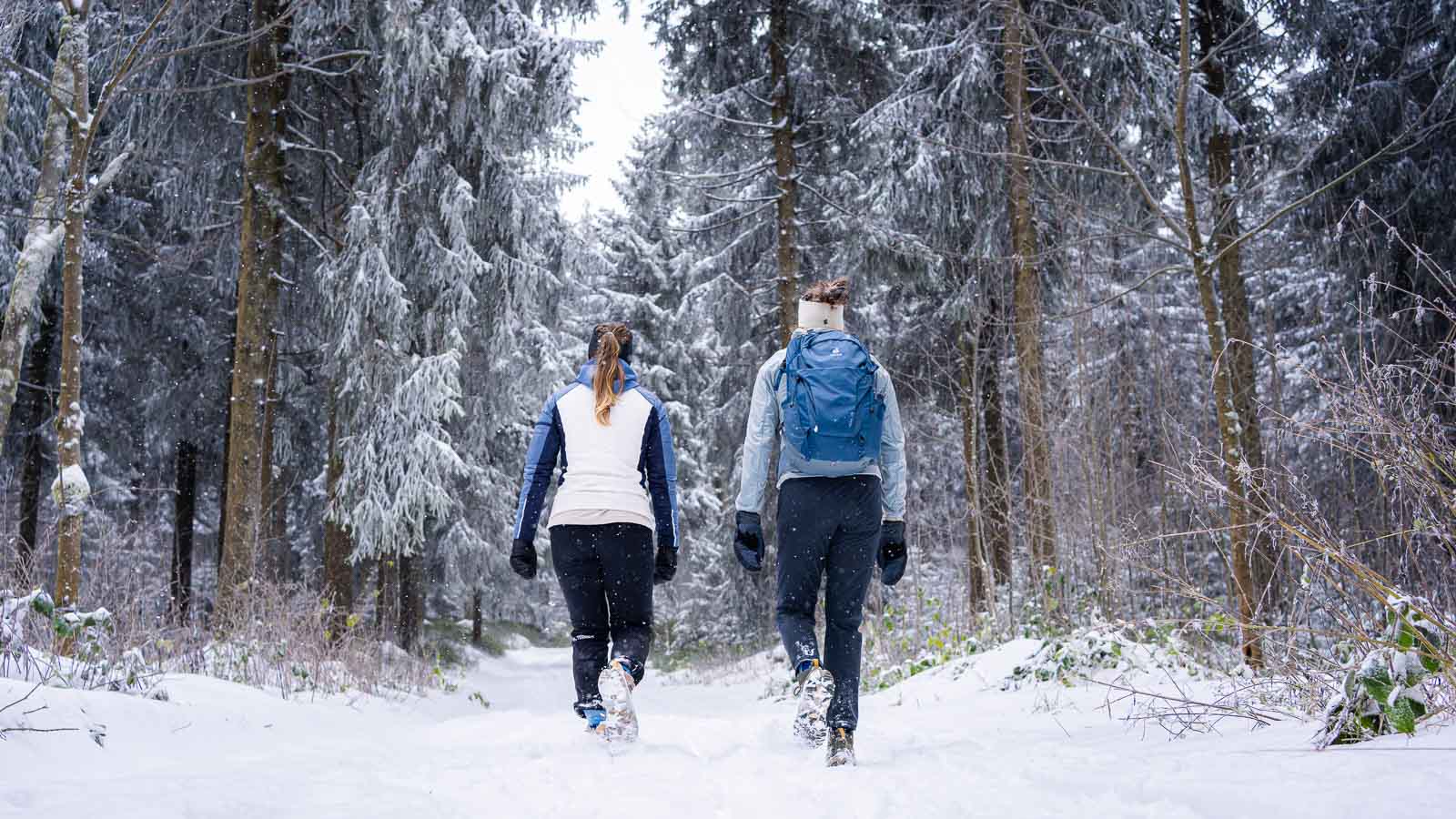 Twee vrouwen lopen door een dik besneeuwd bos, gezien van achteren. Ze dragen winterjassen, wandelschoenen en handschoenen. Een van hen draagt een blauwe rugzak.
