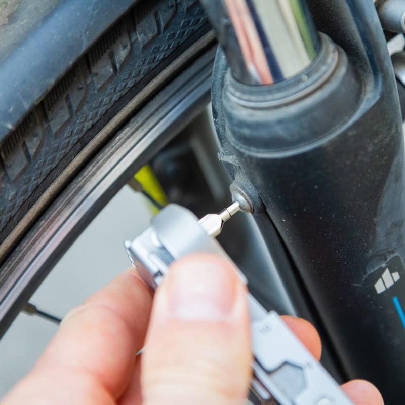 Close-up van een fietsband en voorvork, waarbij een hand met een grijze Bauerfeind Sports multitool een kleine schroef nauwkeurig afstelt – voor betrouwbare techniek en meer zekerheid tijdens elke rit.