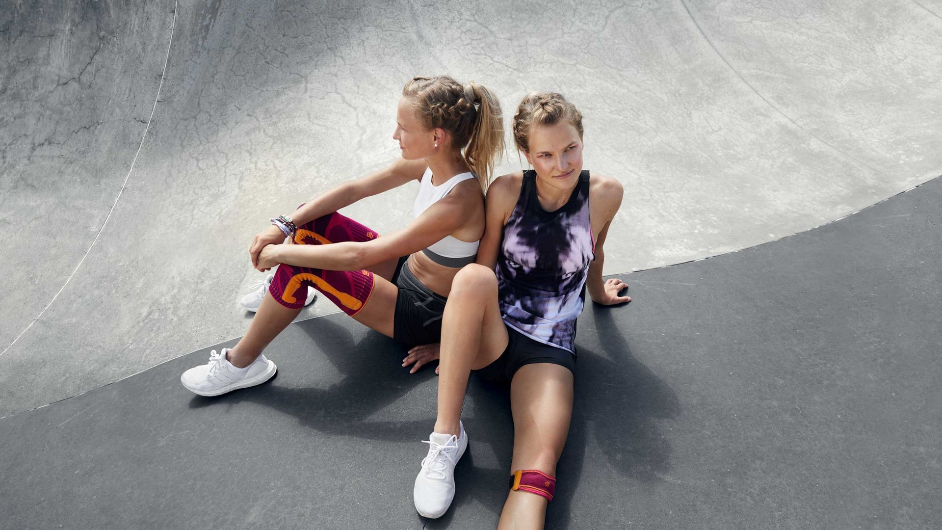 Twee jonge vrouwen in sportkleding zitten rug aan rug op de grond van een skatebaan. Beiden dragen witte sportschoenen, de vrouw links een wit sporttop en kleurige compressiekousen, de vrouw rechts daarnaast een kniebandje.