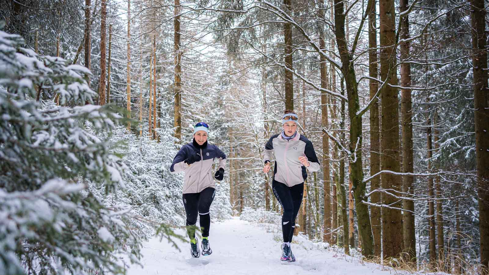Twee hardlopers joggen op een met sneeuw bedekt bospad. De besneeuwde sparren vormen een dicht, winters decor. Beiden dragen warme jassen en hoofdbanden voor een winterse hardlooptocht.