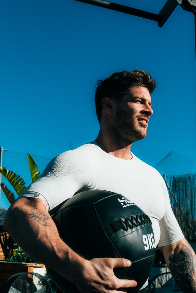 Atleet staat buiten met een zwarte medicinebal van 9 kg in zijn armen, draagt een nauwsluitend sportshirt en kijkt in het zonlicht vooruit – functionele krachttraining in de context van hybrid athlete training.