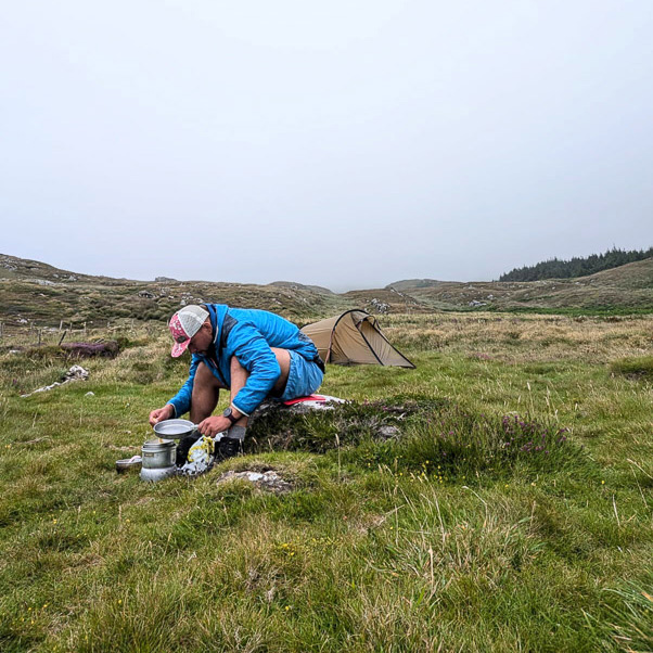Een persoon in een blauwe jas zit op een weide in heuvelachtig terrein voor een kleine tent en bereidt een maaltijd op een campingkooktoestel. De lucht is bewolkt en het landschap oogt ruw en ongerept.