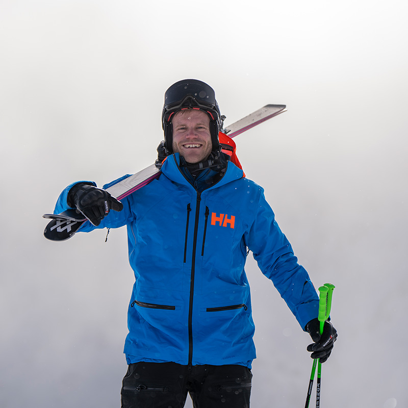Een man staat voor een mistige achtergrond, draagt een blauwe jas en een helm met skibril en heeft zijn ski's op zijn schouder.