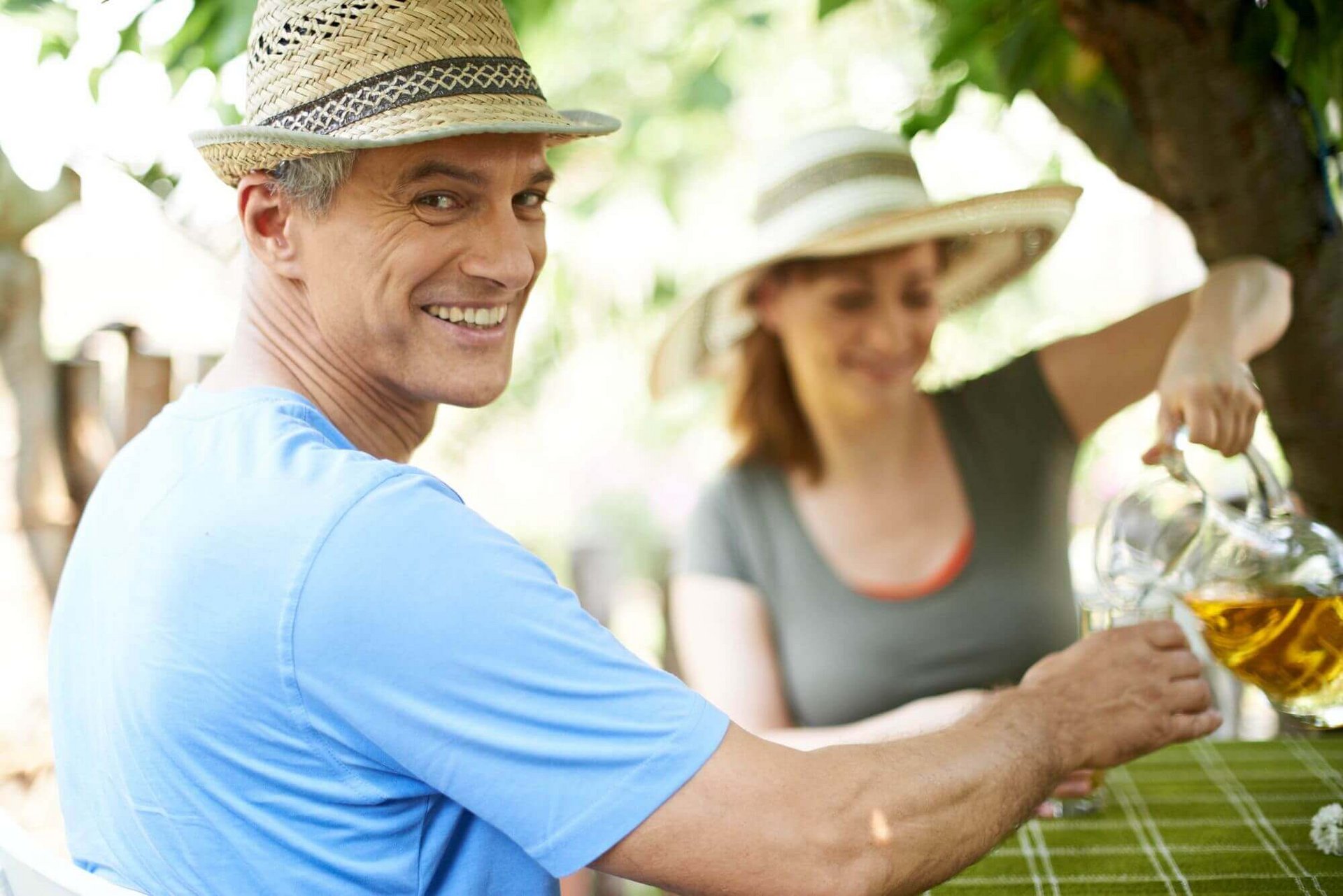 Een man en een vrouw zitten allebei met een strohoed aan een tafel in de tuin. De vrouw schenkt de man een glas appelsap in. De man op de voorgrond lacht vriendelijk in de camera.