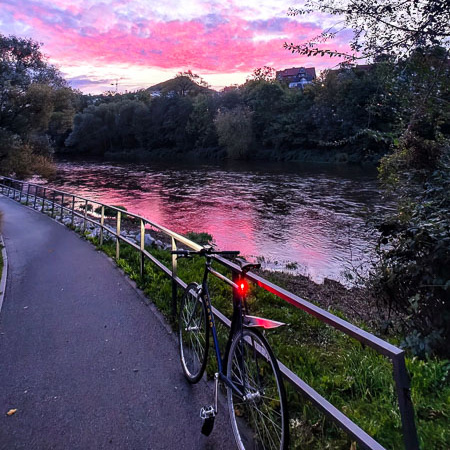 Een fiets leunt tegen een railing langs de rivier, terwijl de lucht paars en roze kleurt. Op de achtergrond weerspiegelen de kleuren in het kalme rivierwater.