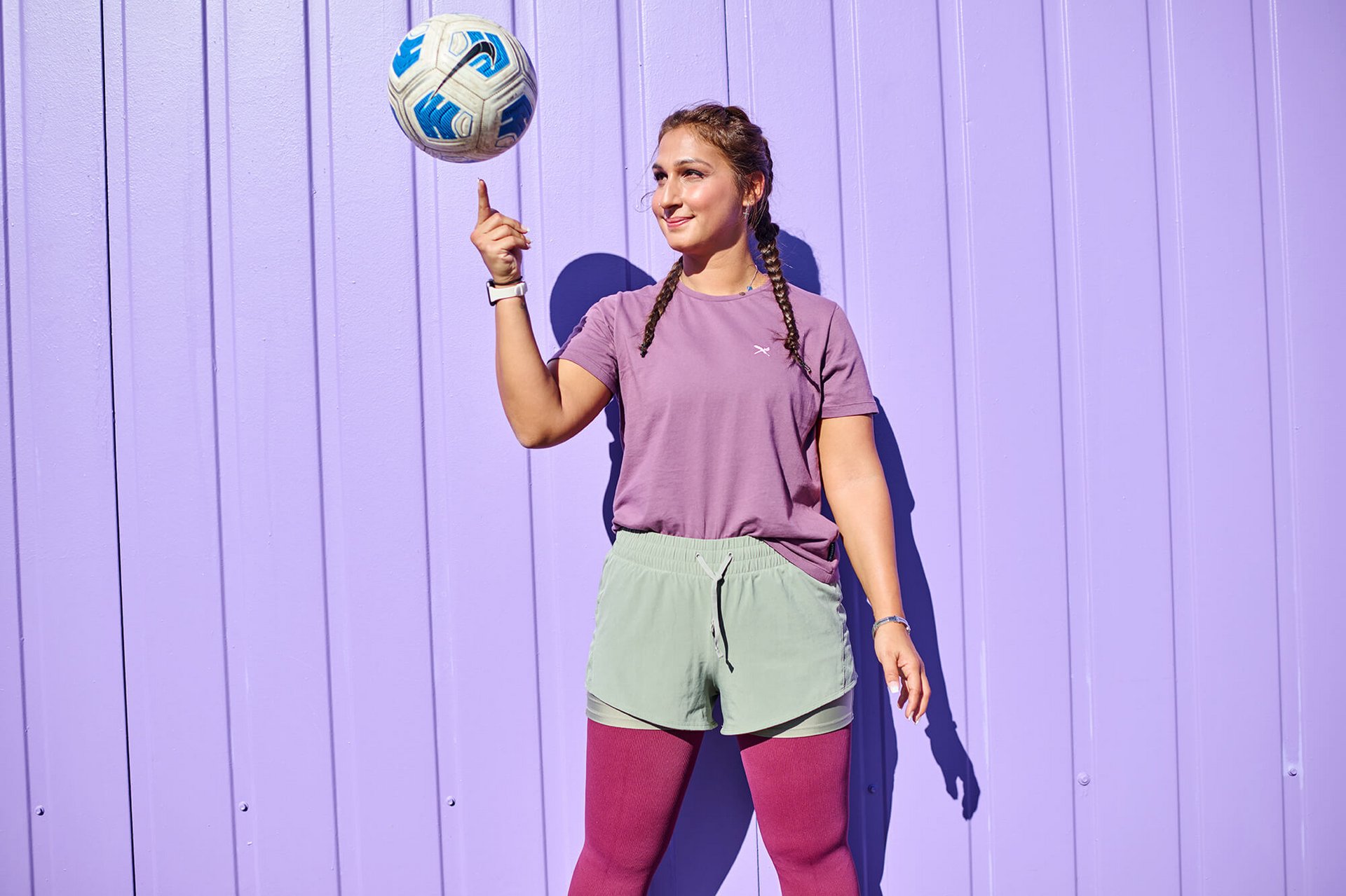 Jonge vrouw in sportieve kleding staat voor een paarse muur en balanceert een bal met blauw patroon op haar vinger. Ze draagt een paars T-shirt, mintgroene short over een bordeauxrode legging en heeft twee gevlochten staartjes.