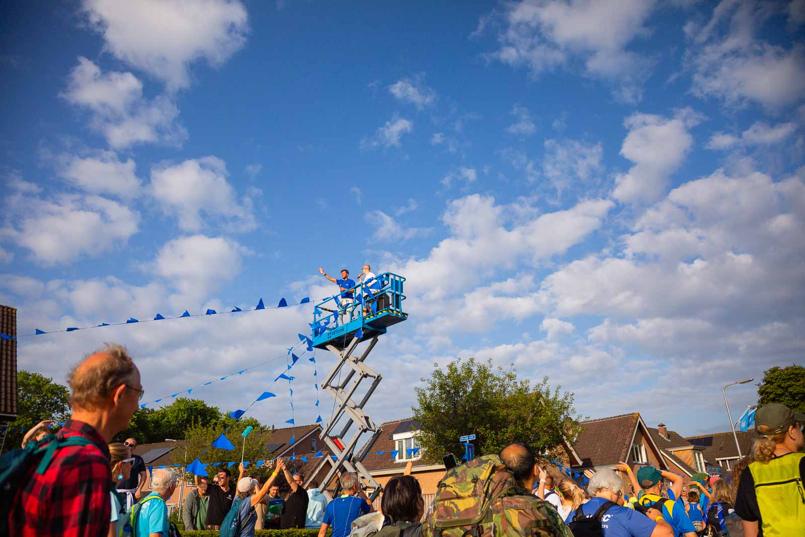 Twee personen zwaaien juichend vanaf een hoogwerker tijdens de 4Daagse in Nijmegen, terwijl wandelaars en toeschouwers onder blauwe vlaggen passeren.