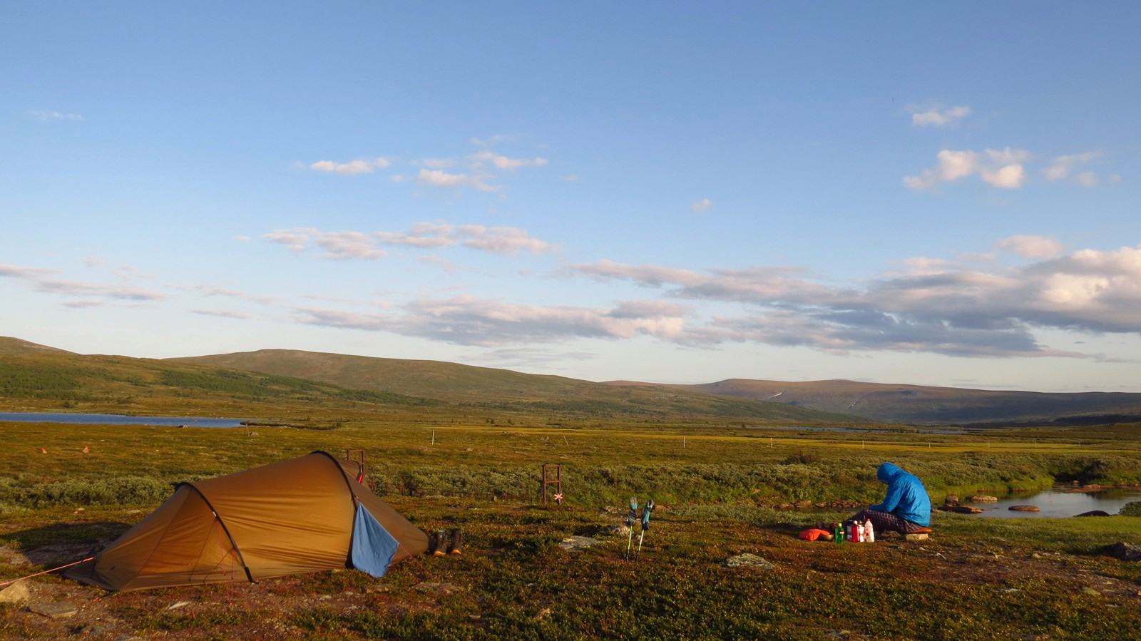Man met een jas met een kap zit voor een tent in een landschap voor een blauwe hemel