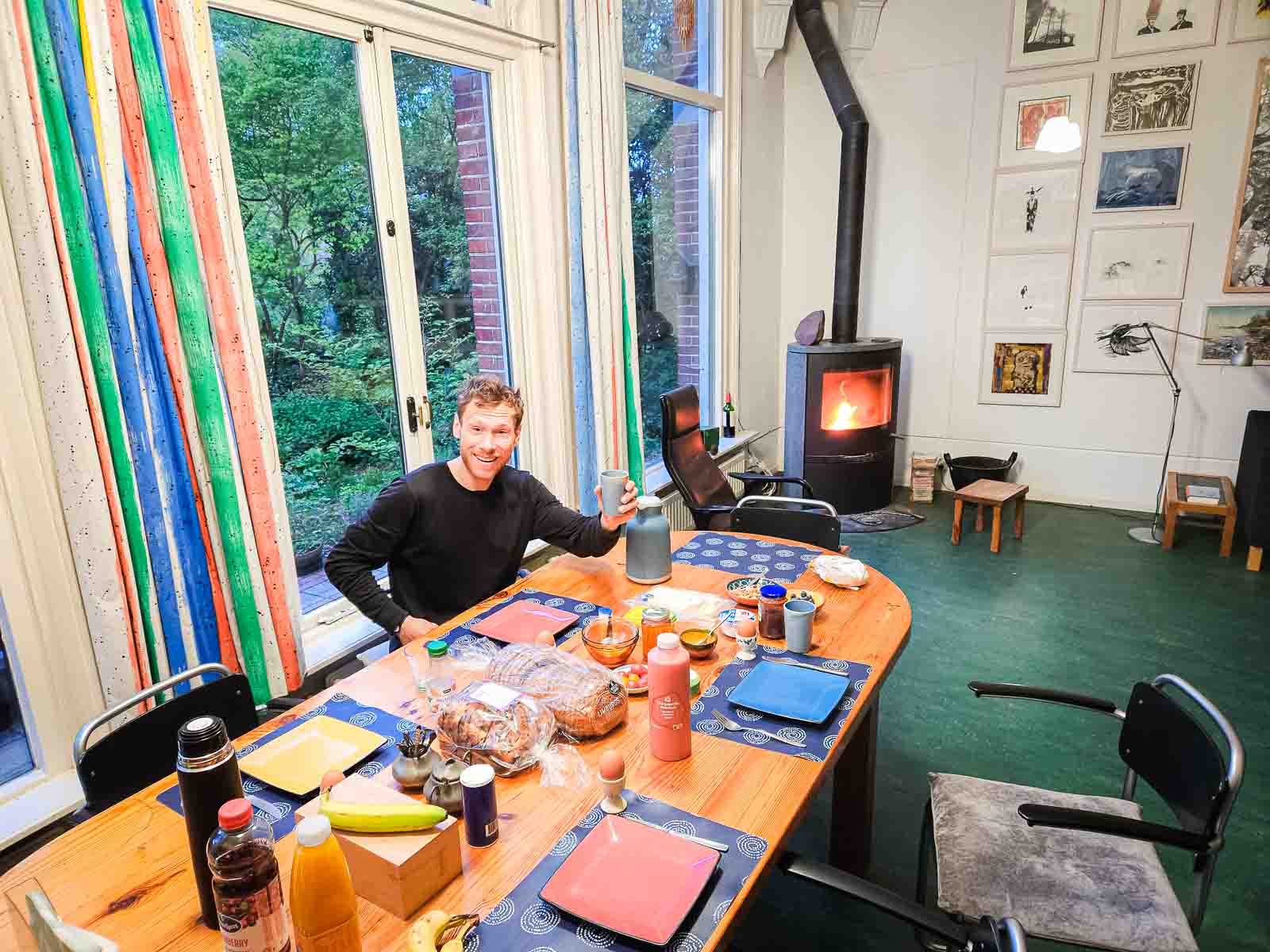 Een man zit aan een rijkelijk gedekte ontbijttafel en proost op de camera.