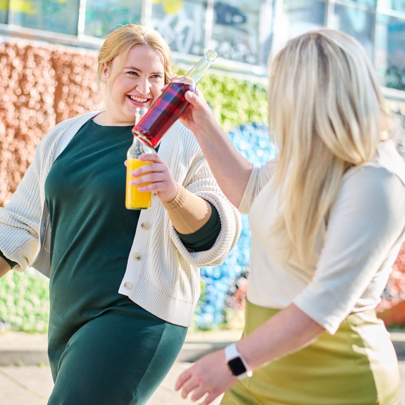 Twee vrouwen klinken met kleurrijke drinkflessen terwijl ze lachend door een stedelijke omgeving wandelen. Op de achtergrond is een kleurrijke muur met grafische patronen te zien, die een levendige sfeer creëert.