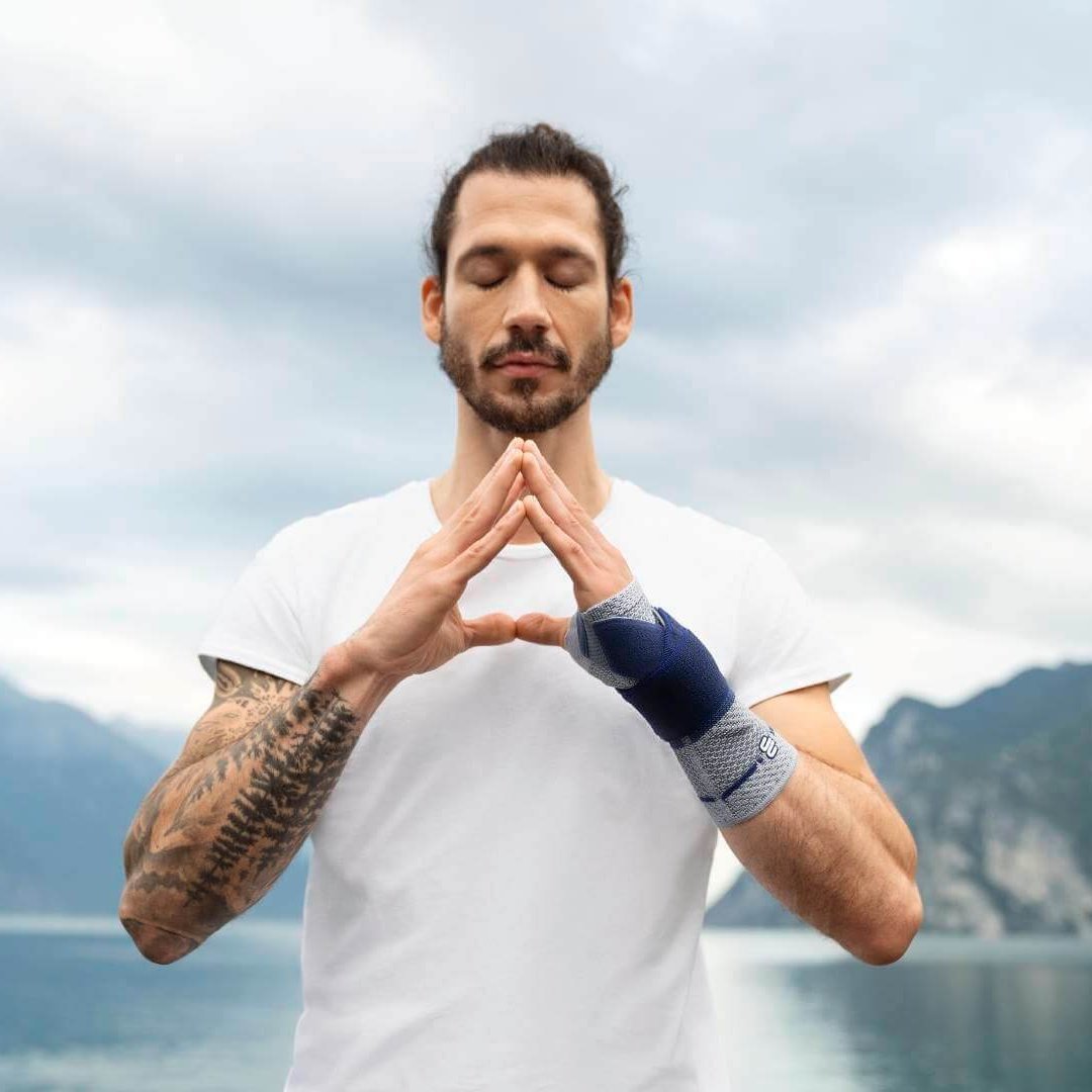 Op de foto is een jonge man bij een meer te zien die een yogahouding aanneemt en daarbij een ManuTrain polsbandage draagt om pijn in de pols te verlichten.
