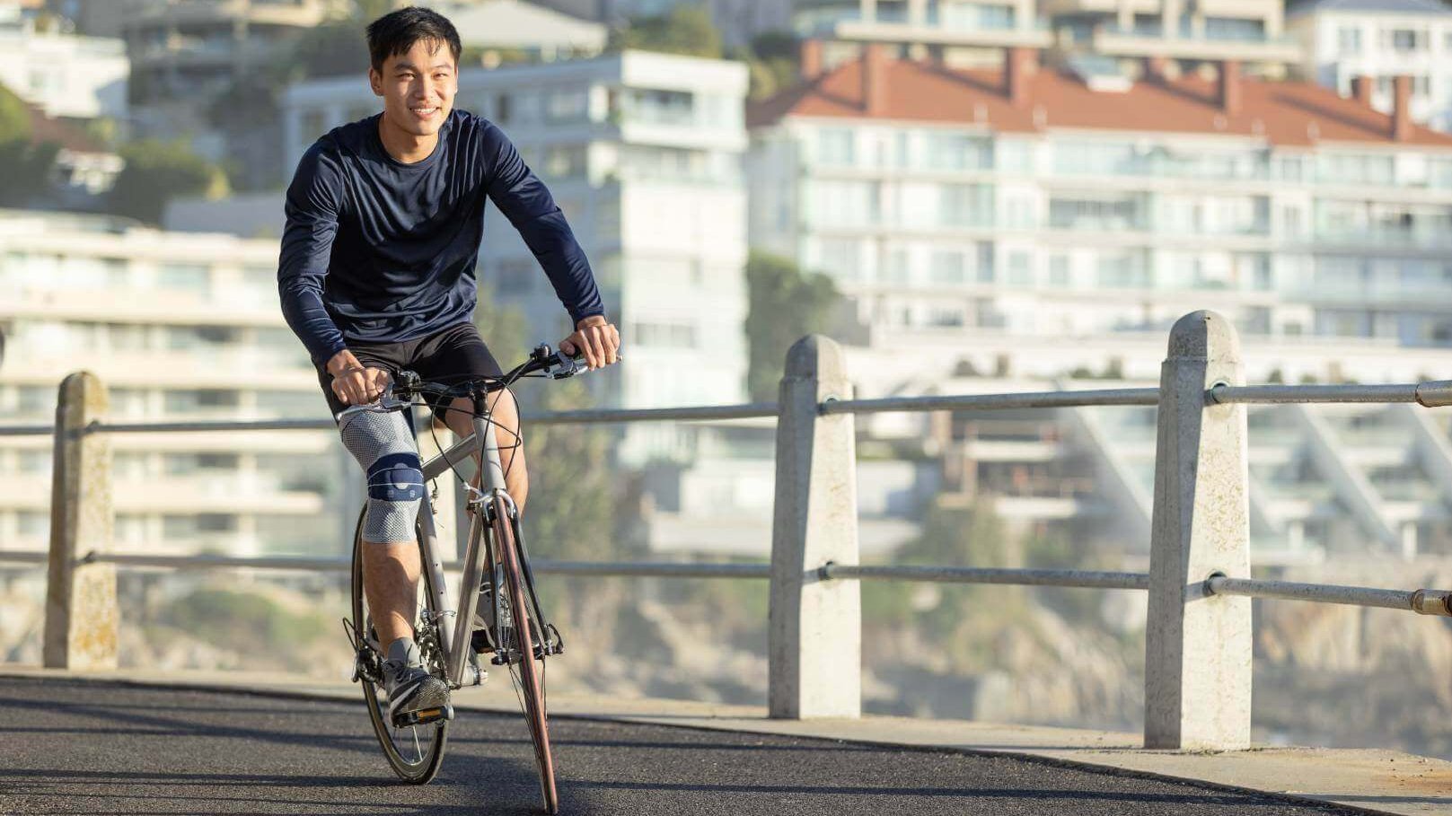 De foto toont een man die met een fiets langs een strandboulevard rijdt en daarbij een GenuTrain bandage draagt om zijn knieklachten te verlichten. Fietsen blijkt een gewrichtsvriendelijke sport.