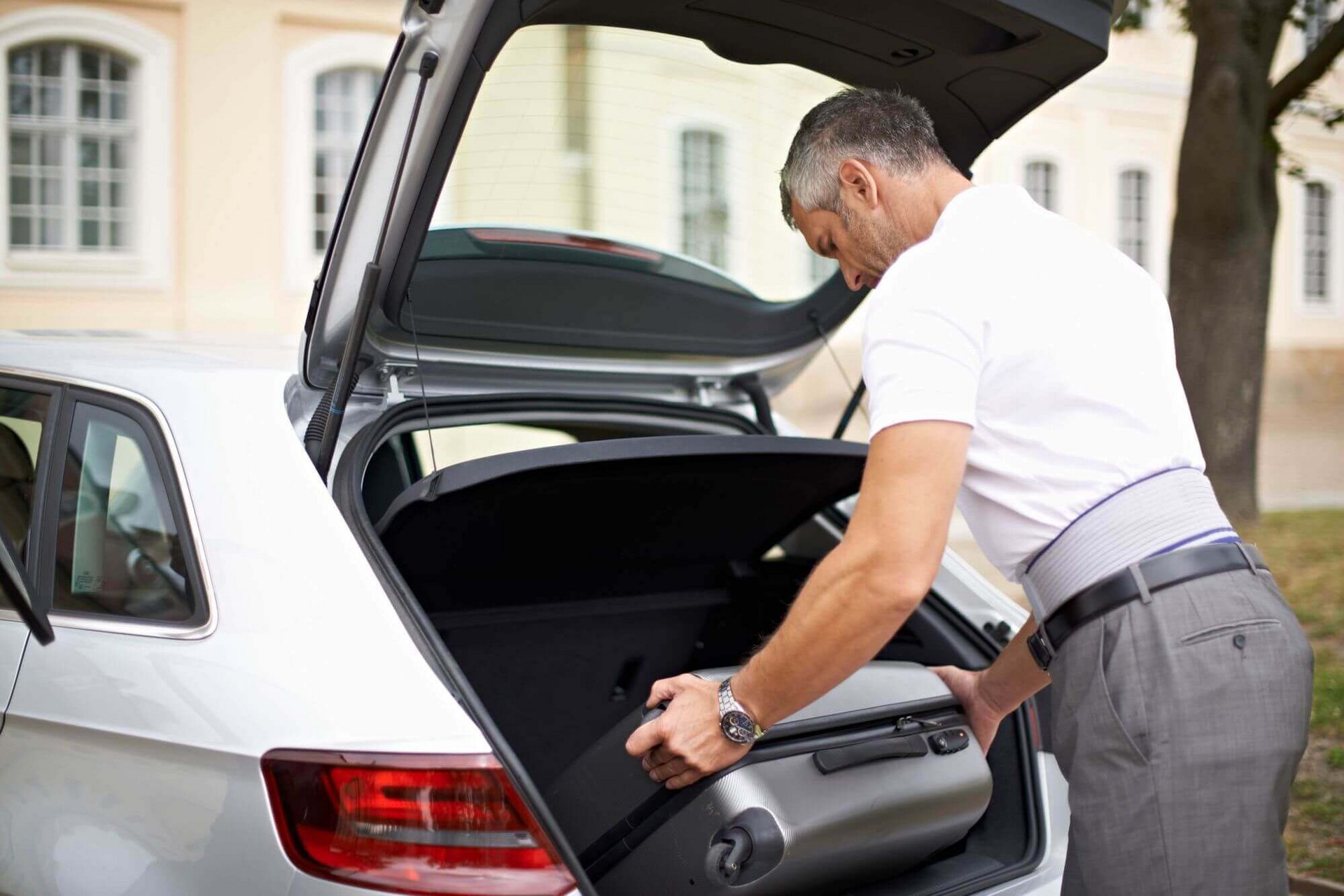 Ein Mann hebt einen Reisekoffer in den Kofferraum eines Autos. Er trägt eine LumboTrain Rückenbandage von Bauerfeind. Rückenbandagen wie die Bauerfeind LumboTrain stabilisieren die Lendenwirbelsäule während und nach einem akuten Lendenwirbelsäulen-Syndrom