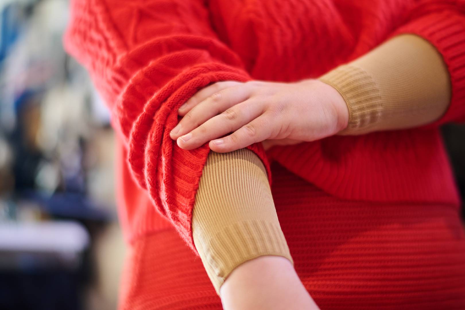 Close-up van een persoon die de mouw van haar rode gebreide trui omhoogtrekt en een beige compressiemouw daaronder laat zien. De scène legt de focus op de ondersteuning door compressiekleding in het dagelijks leven.